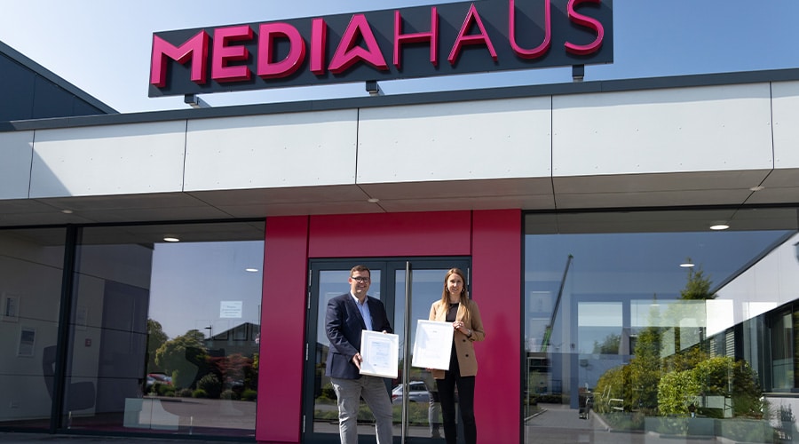 Der Geschäftsführer und eine Mitarbeiterin vom MEDIAHAUS mit der Zertifizierung für eine Top Qualität in der Hand vorm Eingang des MEDIAHAUSES.