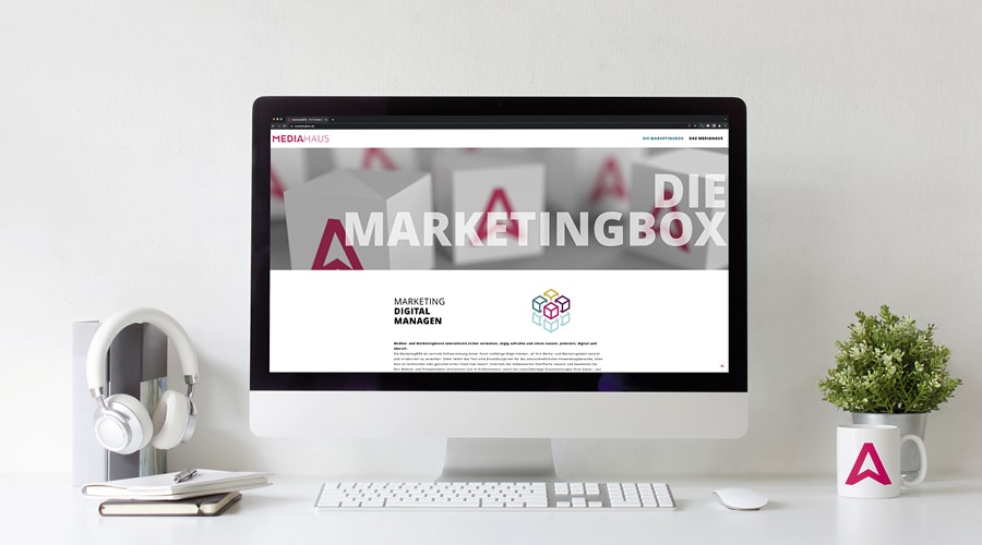 Bild eines Schreibtisches mit PC-Bildschirm dem die Webseite des MEDIAHAUSES zum Thema "Marketingbox" zusehen ist. Desweiteren eine Tastatur, eine MEDIAHAUS-Tasse, eine Blume, Bücher und Kopfhörer.