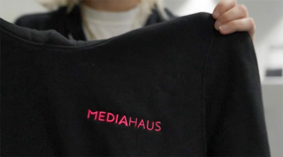 Nicht erkennbare Person hält einen schwarzen Pullover mit der pinken Aufschrift MEDIAHAUS aus dem eigenen Brandstore in die Kamera.